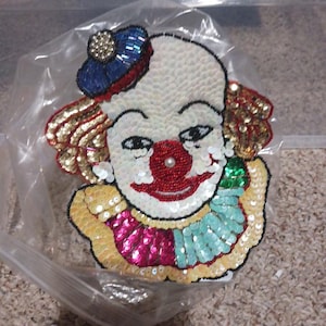 May include: A decorative clown patch featuring intricate beadwork and sequins. The clown has a white face, a red nose with a central pearl, a blue hat, gold hair, and a ruffled collar in alternating sections of yellow, pink, and light green.