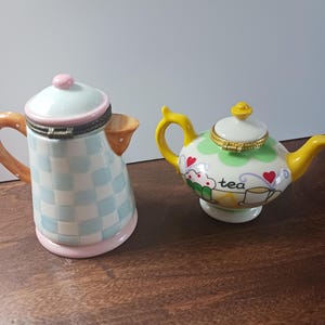 May include: Two decorative ceramic teapots. One is a tall, checkered blue and white design with a pink base and lid. The other is a small, round teapot with a green and white pattern, yellow handle and spout, and the word "tea" on the side.