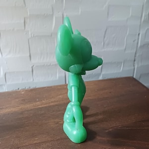 May include: A solid green plastic toy figure of a cartoon mouse, standing on a wooden surface. The figure has large ears, a long nose, and is wearing a shirt and shorts. The toy is a bright, solid green color.
