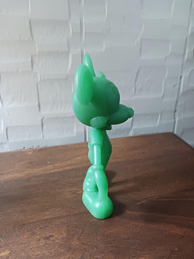 May include: A solid green plastic toy figure of a cartoon mouse, standing on a wooden surface. The figure has large ears, a long nose, and is wearing a shirt and shorts. The toy is a bright, solid green color.