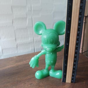 May include: A green Mickey Mouse figurine stands next to a ruler. The figurine is approximately 4 inches tall. The ruler shows measurements in inches. The figurine is on a brown surface.