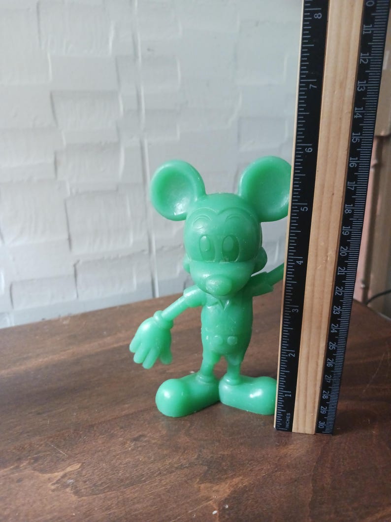 May include: A green Mickey Mouse figurine stands next to a ruler. The figurine is approximately 4 inches tall. The ruler shows measurements in inches. The figurine is on a brown surface.