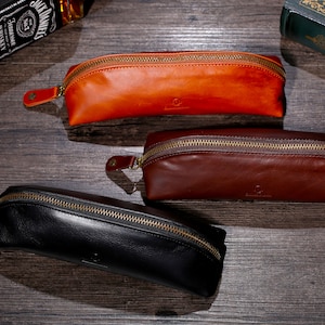 Op de afbeelding: Drie leren potloodkokers in verschillende kleuren: oranje, bruin en zwart. Elke koker heeft een ritssluiting en een subtiel logo.