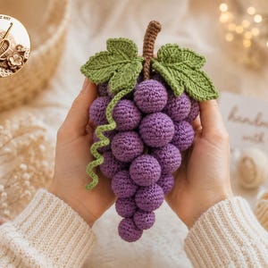 May include: A handmade crochet grape cluster featuring numerous small, round, purple yarn spheres arranged in a bunch. The fruit includes two textured green leaves and a brown stem at the top, with a single curly green vine extending down the side.