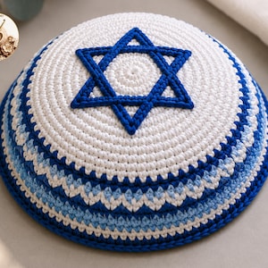 Op de afbeelding: Een gehaakte witte keppel met een opvallende blauwe Davidster in het midden. De basis van het hoofddeksel is versierd met een herhalend zigzagpatroon in afwisselende tinten van donkerblauw, lichtblauw en wit garen met een textuur.
