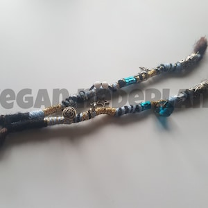 Puede incluir: Dos hebras de hilo azul, negro, blanco y dorado están trenzadas juntas con cuentas y abalorios. Las hebras están atadas en los extremos con cabello castaño.