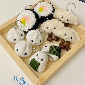 May include: A collection of handmade crochet food keychains, including sushi rolls, dumplings, and rice balls, arranged in a wooden tray. The keychains feature detailed stitching and smiling faces, with silver key rings attached.