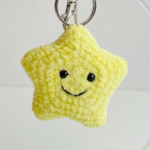 May include: A yellow star-shaped keychain with a smiling face. The plush star has a textured surface and is attached to a silver metal keyring and clasp. The background is a soft white.