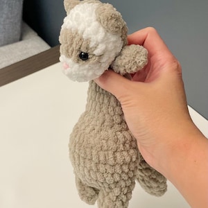 May include: A handmade crochet cat plushie in shades of gray and white. The cat has a fluffy texture, small ears, and a pink nose. The plushie is held in a hand, showcasing its size and detail. The overall theme is handmade toys.