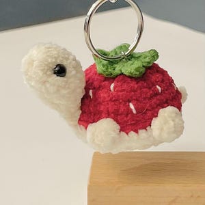May include: A crocheted strawberry turtle keyring. The turtle has a red shell with white detailing, a green leafy top, and a white body. It is attached to a silver keyring.