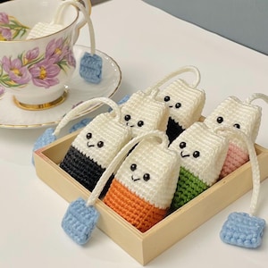 Puede incluir: Una bandeja de madera contiene varias bolsitas de té de ganchillo con caras sonrientes y fondos coloridos. Cada bolsa tiene una parte superior blanca y una etiqueta de ganchillo azul. Una taza de té con diseño floral está en el fondo.