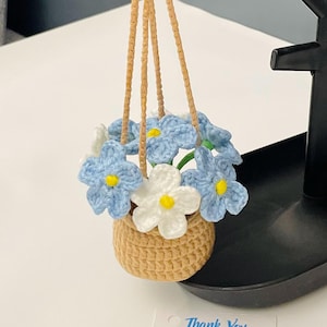May include: A crocheted miniature basket filled with blue and white flowers with yellow centers, suspended by tan cords. The basket is tan, and the flowers are arranged to appear full. A small tag with text is visible.