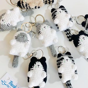 May include: Handmade crochet cat keyrings in various colours, including white, grey, black, and tan. Each keyring features a cat-shaped design with a metal keyring and clasp. The keyrings are displayed on a white surface.