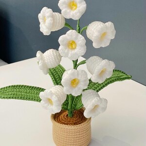 Puede incluir: Un arreglo floral de ganchillo hecho a mano con flores blancas en forma de campana con centros amarillos, hojas verdes y un tallo marrón, todo en una maceta de ganchillo beige. Una pieza decorativa encantadora.