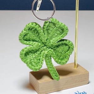 May include: A handmade green crochet four-leaf clover keychain. The clover is attached to a silver keyring. The keychain is displayed on a wooden stand with a gold-colored metal support. A white card with blue text is in the background.