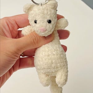 May include: A white plush cat keychain with a silver metal ring. The cat has black eyes and a pink nose. The keychain is made of a soft, textured material.