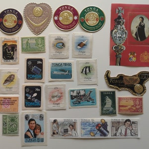 Op de afbeelding: Een verzameling postzegels uit Tonga, met verschillende ontwerpen, waaronder een satelliet, een vliegtuig, een vis en een koninklijk paar. De postzegels zijn gerangschikt in rijen en kolommen op een witte achtergrond.