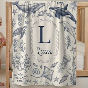 May include: A cream-colored baby blanket featuring a navy blue nautical print of sea creatures including a whale, orca, dolphin, octopus, sea turtle, seahorse, and jellyfish. A central rope-style oval frame contains the letter L and the name Liam.