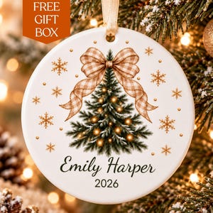 May include: A round white ceramic ornament featuring a green Christmas tree with glowing lights, topped with a tan and white gingham bow. The design includes gold snowflakes and the text Emily Harper 2026 printed in black script and sans-serif font.