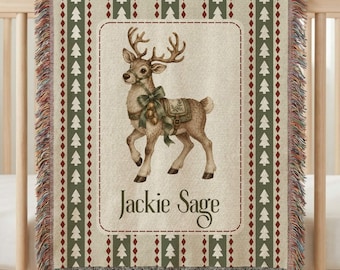 May include: A woven throw blanket featuring a central illustration of a reindeer wearing a green bow and a saddle. The blanket has a cream background with a border of green pine trees and red diamonds. The name Jackie Sage is printed below.