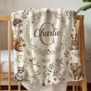 May include: A cream-colored fleece blanket featuring a central wreath design surrounding the name Charlie and a large letter C. The blanket is decorated with illustrations of a hedgehog, squirrel, rabbit, badger, raccoon, bear, and birds.