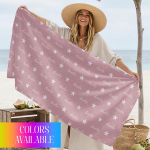 May include: A dusty rose beach towel with a repeating pattern of white stars and the name "Amelia" in a script font. The towel is rectangular and held up by a person. The words "COLORS AVAILABLE" are in a colorful box.