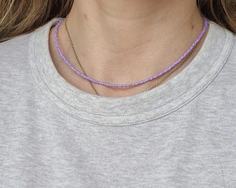 Könnte beinhalten: Eine zarte, hellviolette Perlenkette. Die Halskette besteht aus kleinen, gleichmäßig verteilten Perlen, die einen subtilen, eleganten Look erzeugen. Eine dünnere Goldkette ist ebenfalls sichtbar. Die Halskette wird auf einem grauen T-Shirt getragen.