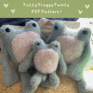 May include: A family of three fuzzy, crocheted frogs in shades of blue and pink. The frogs are sitting on a light brown surface. The largest frog is on the right, the middle-sized frog is on the left, and the smallest frog is in the centre. The image includes the text "Fuzzy Froggy Family PDF Pattern!"