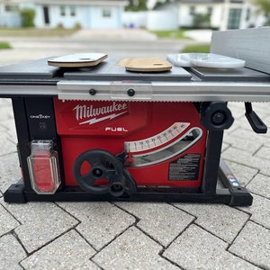 May include: A red Milwaukee FUEL table saw with a black base and a silver sliding table. The saw has a clear plastic dust collection container and a black knob for adjusting the blade height. The saw is on a gray brick surface.