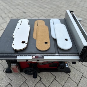 Peut inclure: Une scie à table avec un plateau noir et une clôture argentée. Il y a quatre plaques de levage de toupie sur la table, deux blanches et deux en bois.