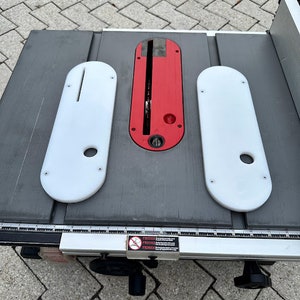 May include: A table saw with three white plastic inserts and a red insert with a slot in the center. The table saw has a ruler with markings in inches.