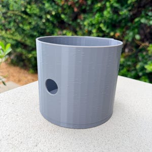 May include: A cylindrical gray container with a smooth, vertical-ribbed texture and a single circular opening on the side. The object features a hollow interior and a flat base, constructed with visible 3D printed layer lines throughout the surface.