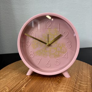 Puede incluir: Un reloj analógico rosa con manecillas doradas y un diseño floral dorado en el centro. El reloj tiene una esfera transparente y está sobre una superficie de madera. El diámetro del reloj es de aproximadamente 10 cm.