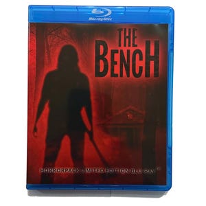 May include: A blue Blu-ray disc case with a red cover. The cover displays the title "THE BENCH" in large, black letters. A silhouetted figure with a weapon is in front of a forest and house. The text "HORRORPACK LIMITED EDITION BLU RAY" is at the bottom.