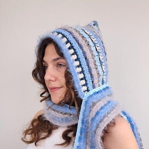 Könnte beinhalten: Ein blauer und grauer Häkel-Kapuzen-Schal mit einer flauschigen Textur. Der Schal hat ein gestreiftes Muster und wird von einer Person mit langen braunen Haaren getragen.