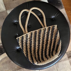 May include: A beige and black striped crochet tote bag with matching handles. The bag is made of a woven material and has a rounded shape. The bag is sitting on a black leather ottoman.