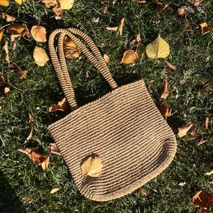 May include: A brown woven tote bag with thick straps. The bag is made of a natural material and has a slightly textured surface. The bag is sitting on a bed of dry leaves.