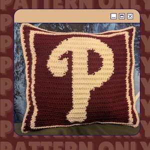 May include: Crocheted pillow with a maroon background and a cream-colored "P" in the center. The pillow has a cream-colored border and a maroon frame. The pillow is square and appears to be handmade.