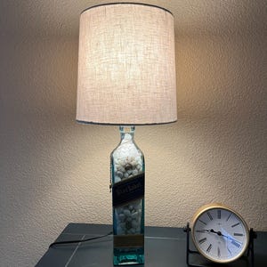 May include: A lamp made from a repurposed clear glass bottle with a white label that says "Blue Label" and white stones inside. The lamp has a beige linen lampshade and is sitting on a dark surface next to a gold clock.