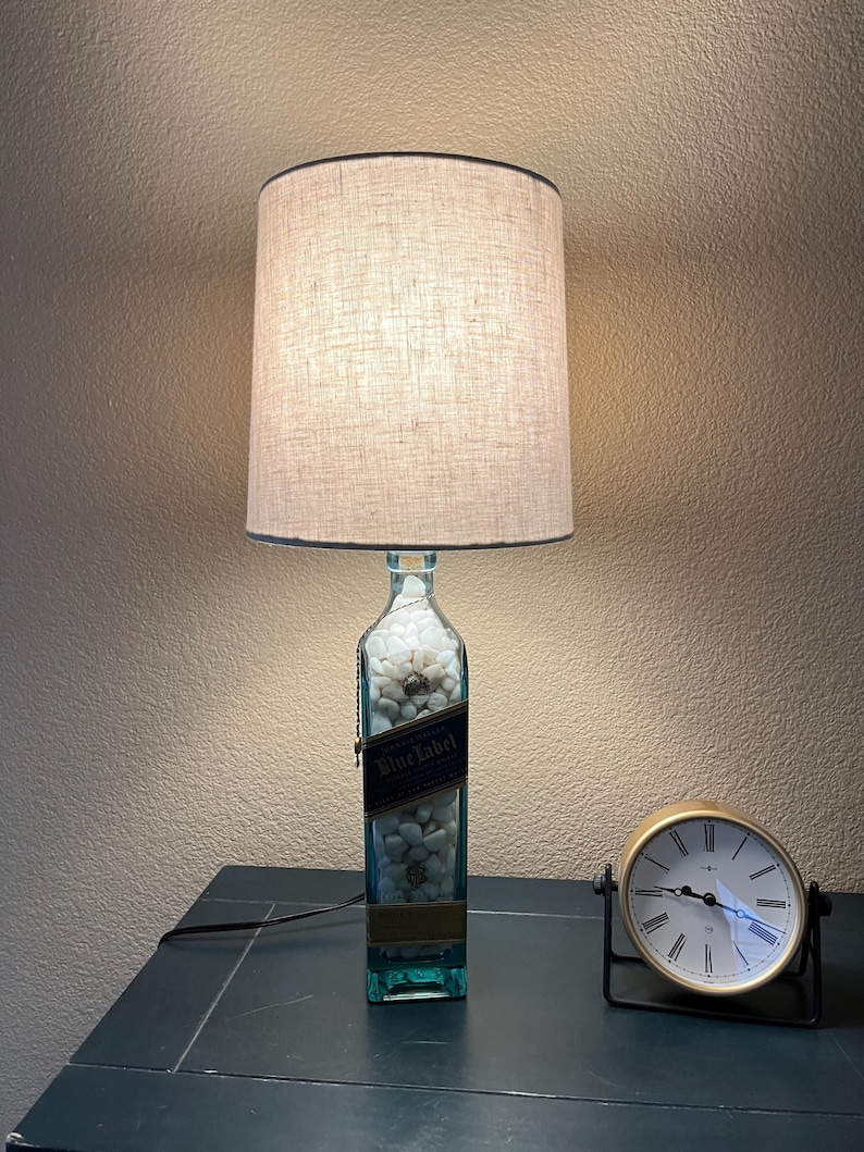 May include: A lamp made from a repurposed clear glass bottle with a white label that says "Blue Label" and white stones inside. The lamp has a beige linen lampshade and is sitting on a dark surface next to a gold clock.