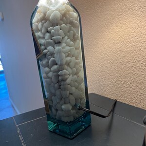 May include: A clear glass bottle lamp with white rocks inside. The bottle is blue at the bottom and has a black cord.