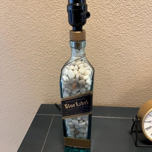 May include: A repurposed Johnnie Walker Blue Label bottle lamp with a black lampshade and white rocks inside. The bottle is clear glass with a blue label and gold lettering.
