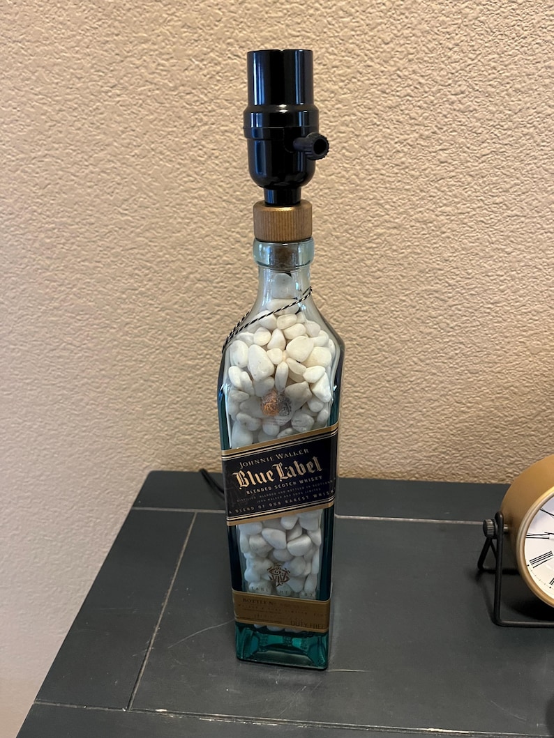 May include: A repurposed Johnnie Walker Blue Label bottle lamp with a black lampshade and white rocks inside. The bottle is clear glass with a blue label and gold lettering.