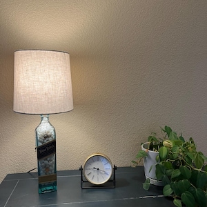 May include: A lamp made from a repurposed bottle with a white fabric shade. The bottle has a label that says "Blue Label" and is filled with white rocks. A gold clock and a potted plant are on a dark wooden surface.
