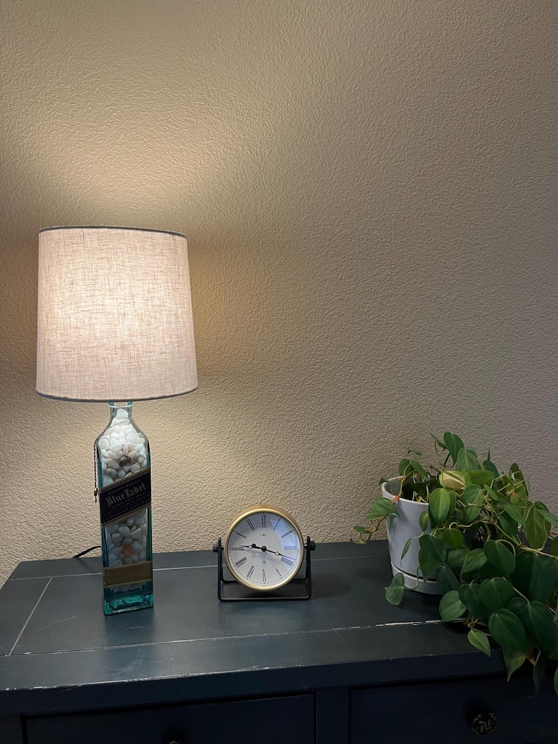 May include: A lamp made from a repurposed bottle with a white fabric shade. The bottle has a label that says "Blue Label" and is filled with white rocks. A gold clock and a potted plant are on a dark wooden surface.