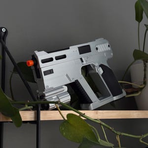 Peut inclure: Un pistolet jouet argenté et noir avec un embout orange repose sur une étagère en bois. L'arme a un design futuriste avec une poignée texturée et plusieurs ouvertures rectangulaires. Des plantes vertes entourent l'arme.