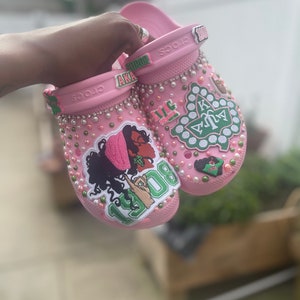 Op de afbeelding: Roze Crocs met strass en groene en roze patches. De patches bevatten de letters AKA, het jaar 1908 en een groen Grieks letterontwerp.