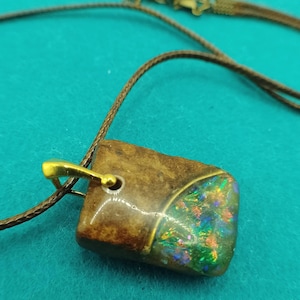 Könnte beinhalten: Eine quadratische Anhängerkette mit einem braunen Stein und einer goldfarbenen Öse. Der Stein hat einen schillernden Bereich mit grünen, blauen und orangen Farbtönen. Die Halskette ist braun.