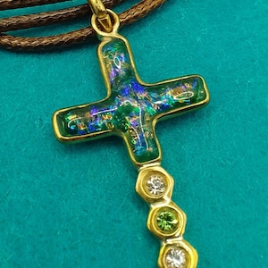 Puede incluir: Un colgante de cruz de oro con una incrustación de vidrio verde y azul iridiscente. El colgante está unido a un collar de cordón marrón y presenta una fila de pequeñas piedras preciosas transparentes y verdes.