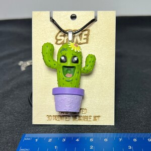 May include: A green 3D printed cactus wearing a purple pot with a white flower on top. The cactus is smiling and has big eyes. The pendant is hanging from a black cord and is labeled "STIKE 3D PRINTED WEARABLE ART".